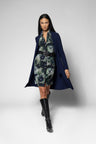 A model walks against a light gray studio background wearing a navy blue open-front coat over a green and gray floral print dress with a black belt, paired with black knee-high boots.