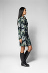 A woman in profile wearing a long-sleeve collared shirt dress with a green, gray, and black abstract floral pattern, paired with black knee-high boots, against a light gray studio background.