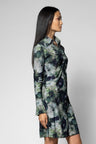 A woman modeling a long-sleeve button-front shirt dress with a green, gray, and navy floral watercolor print, shown from a side profile against a light gray studio background.