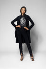 A model wearing a black long-sleeve jacket, black pants, and a black top with a white graphic print, standing against a light gray studio background.