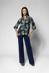 A woman modeling a 3/4 sleeve shirt with a green and black abstract floral pattern, paired with navy blue wide-leg trousers, against a light gray studio background.
