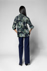 A woman is shown from the back wearing a dark green and navy floral patterned blouse with navy blue trousers, standing against a light gray studio background.