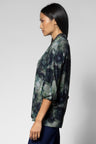 A woman shown in side profile wearing a loose blouse with a dark abstract floral print in navy, green, and gray tones, paired with dark navy bottoms, against a light gray studio background.