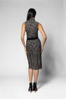 A woman viewed from the back wearing a sleeveless high-neck shirt and matching printed skirt with a textured black and gray pattern, a black waistband, and black ankle boots, set against a light gray studio background.