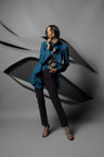 A woman posing in a studio setting wearing a teal blue wrap-style jacket, dark trousers, and patterned sandals, set against a gray background with abstract black shapes.