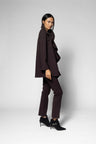A model is shown in profile wearing a dark brown ruffled cape-style jacket, matching dark brown trousers, and black ankle boots against a light gray studio background.