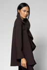 A model wearing a dark brown, draped, ruffled coat with a high collar, photographed against a neutral light gray studio background.