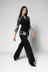 A woman modeling a black long-sleeve top with green and blue abstract patterned sleeves and hem, paired with black wide-leg pants, against a light gray studio background.