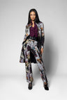 A model stands against a light gray studio background wearing a matching floral print blazer and trousers set with a dark purple ruffled blouse and black shoes.