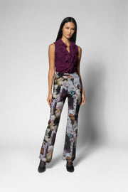 A model stands against a light gray studio background wearing a sleeveless deep purple ruffled top and high-waisted wide-leg trousers with a muted floral and abstract print in gray, purple, beige, and brown tones.