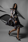 A woman models a long-sleeved, knee-length floral print wrap or coat over a black dress, paired with black pointed-toe heels, set against a stylized gray background with abstract layered paper-like shapes.