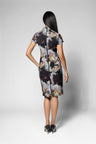 A woman shown from the back wearing a short-sleeve knee-length dress with a dark abstract floral print, photographed against a light gray studio background.