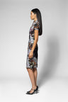 A woman in profile wearing a short-sleeved, knee-length dress with a dark base and a painterly floral pattern in muted gray, brown, and white tones, paired with black pointed-toe heels, against a light gray studio background.