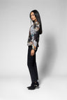 A woman stands in profile wearing a long-sleeve abstract print jacket with black pants and black ankle boots against a light gray studio background.