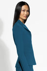 A woman with long, straight black hair is shown in profile wearing a tailored teal-blue blazer against a clean white studio background.