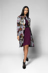 A model is shown wearing a long-sleeved, knee-length floral print coat over a dark purple dress with a black belt, paired with black ankle boots, against a light gray studio background.