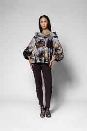 A model stands against a light gray studio background wearing a loose, oversized floral-patterned top with dramatic long sleeves, dark brown slim pants, and strappy high-heeled sandals.