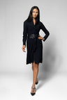 A woman modeling a black coat with long sleeves, a pointed collar, and a wide black belt with a silver buckle, paired with black pointed-toe heels, against a light gray studio background.