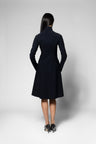 A woman viewed from the back wearing a black long-sleeve knee-length coat with a high collar and hands in pockets, standing against a light gray studio background.