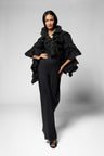 A model wearing a black ruffled top, wrap, and matching wide-leg trousers, standing against a light gray studio background.