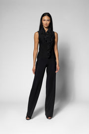 A model stands against a light gray studio background wearing a black sleeveless top with a ruffled collar and matching black wide-leg pants.