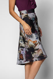 A woman wearing a high-waisted A-line midi skirt with a large abstract floral print in shades of gray, white, black, and brown, paired with a dark waistband and a dark purple top, shown against a light gray studio background.