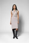 A model stands against a light gray studio background wearing a sleeveless light beige dress with a ruffled tie-neck bodice and a fitted knee-length skirt, paired with black knee-high boots.