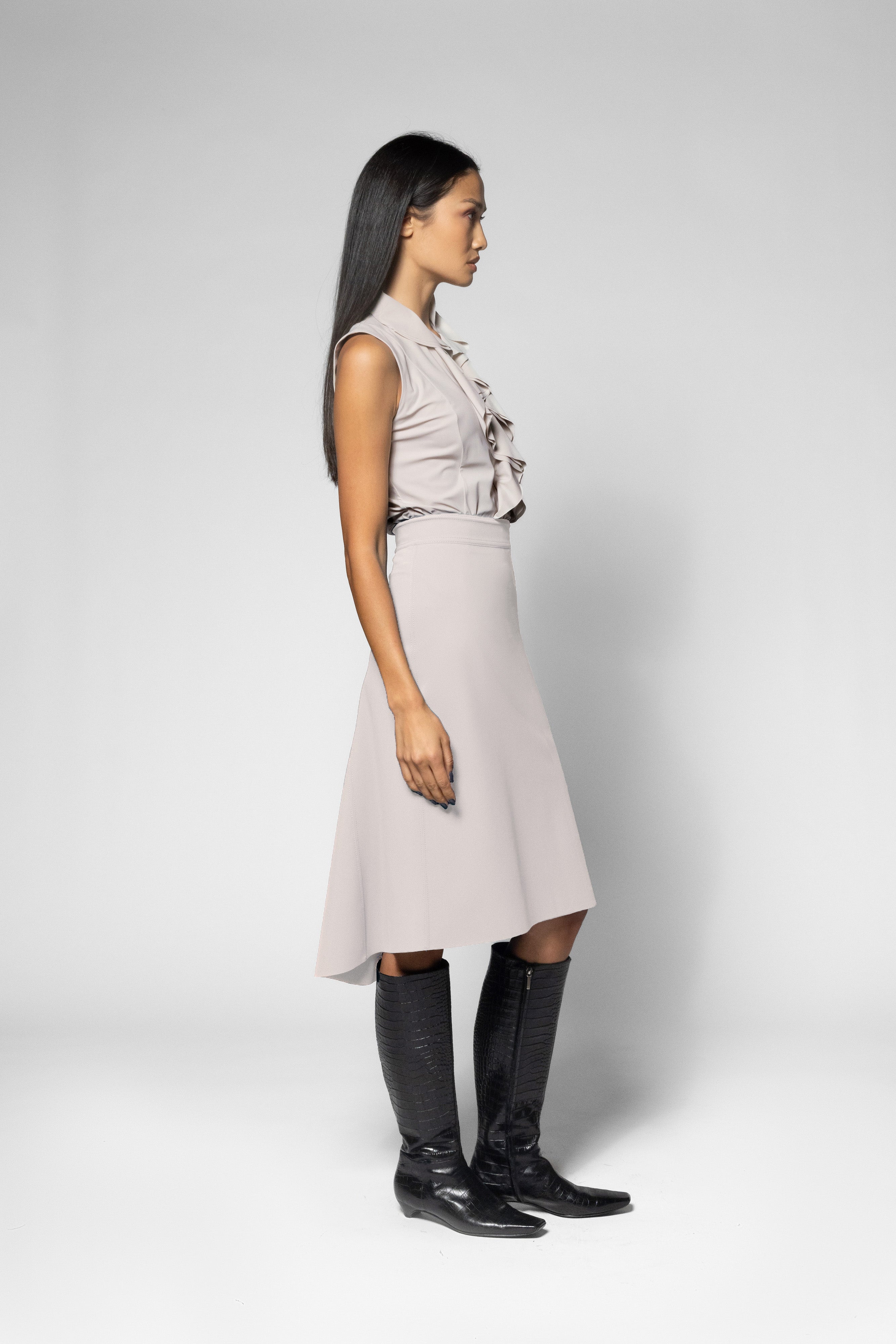 A woman in profile wearing a sleeveless light beige dress with a ruffled collar and waist, paired with black knee-high boots, against a light gray studio background.