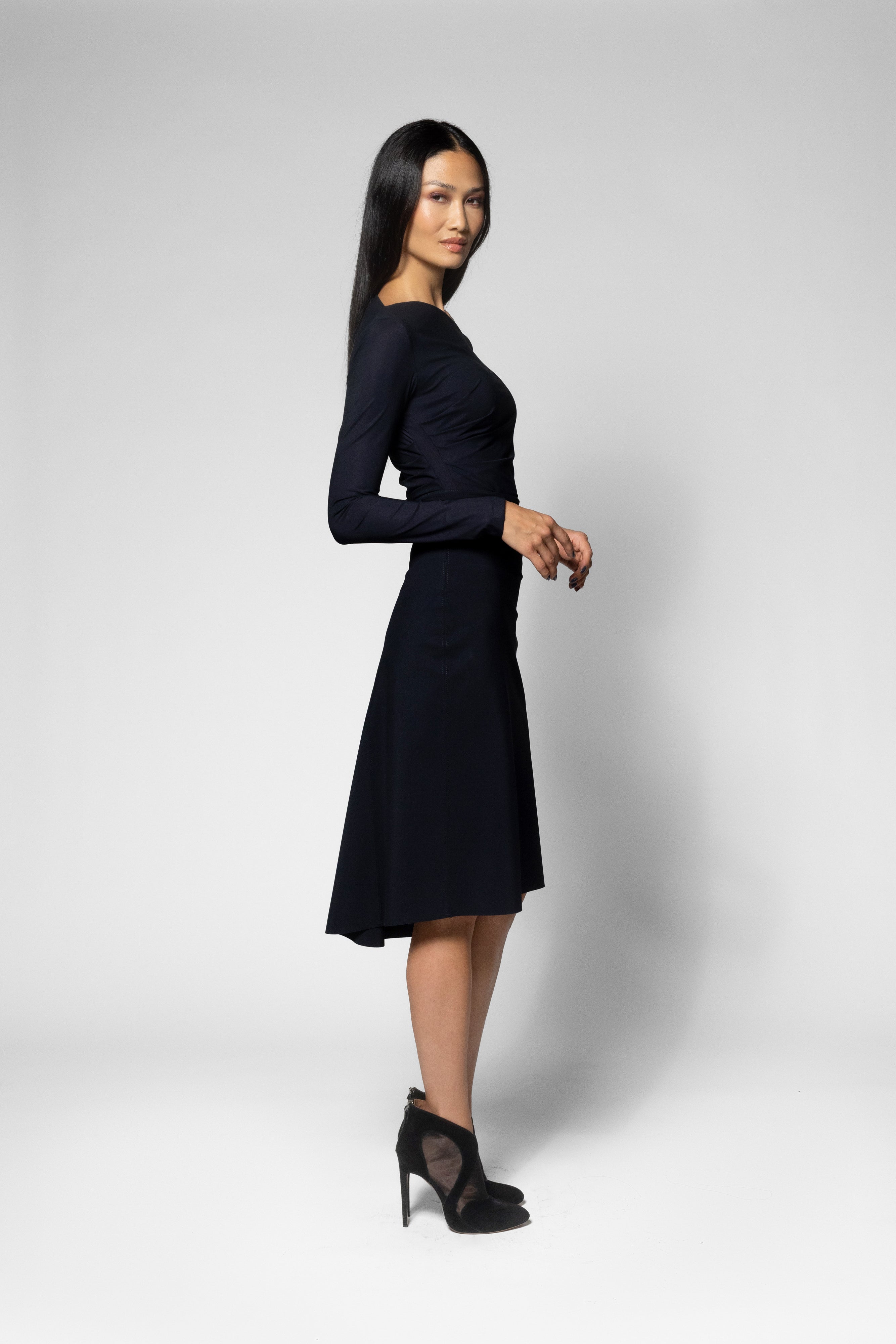 A woman modeling a long-sleeve black shirt and black flared skirt, paired with black high heels, against a light gray studio background.