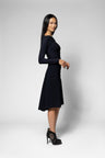 A woman modeling a long-sleeve black shirt and black flared skirt, paired with black high heels, against a light gray studio background.