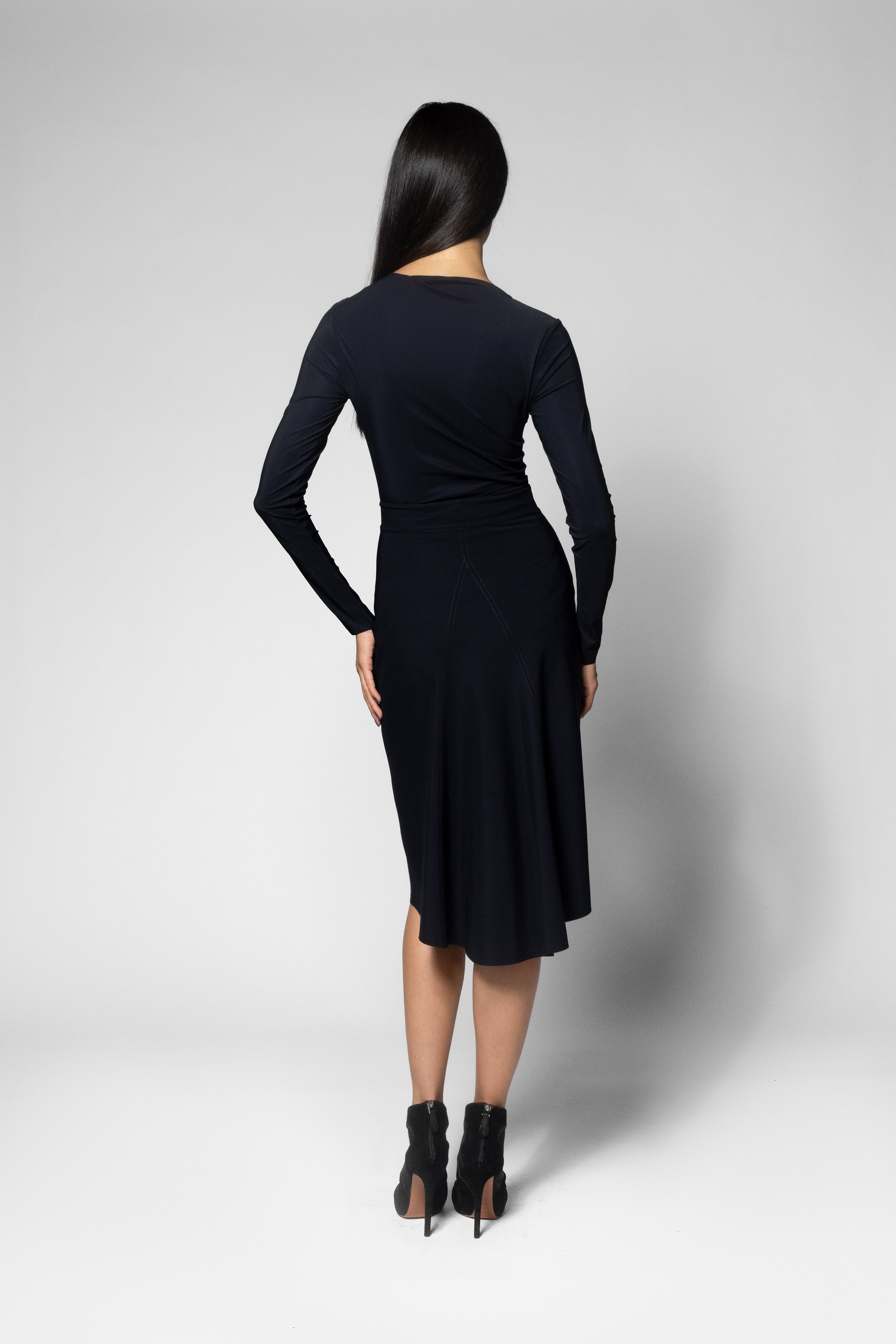 A woman viewed from the back wearing a black long-sleeve shirt and skirt with black ankle boots, standing against a light gray studio background.