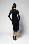 A woman viewed from the back wearing a black long-sleeve shirt and skirt with black ankle boots, standing against a light gray studio background.