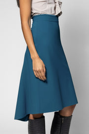 A woman wearing a teal blue A-line midi skirt with a high-low hem, paired with a light beige top and black boots, shown from the waist down against a neutral gray background.