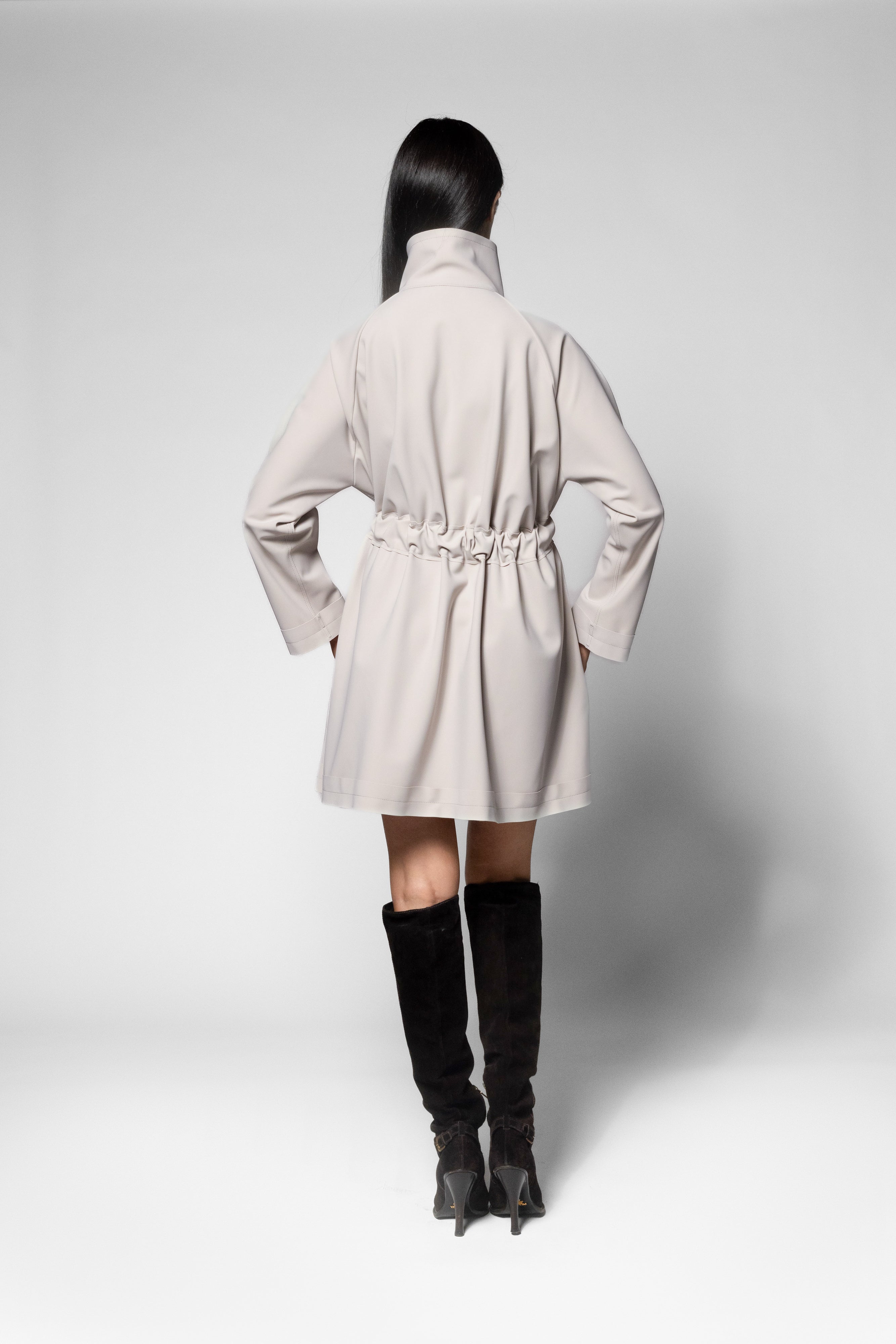 A model shown from the back wearing a light beige trench coat with a drawstring waist and black knee-high boots, set against a plain light gray studio background.