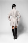 A model shown from the back wearing a light beige trench coat with a drawstring waist and black knee-high boots, set against a plain light gray studio background.