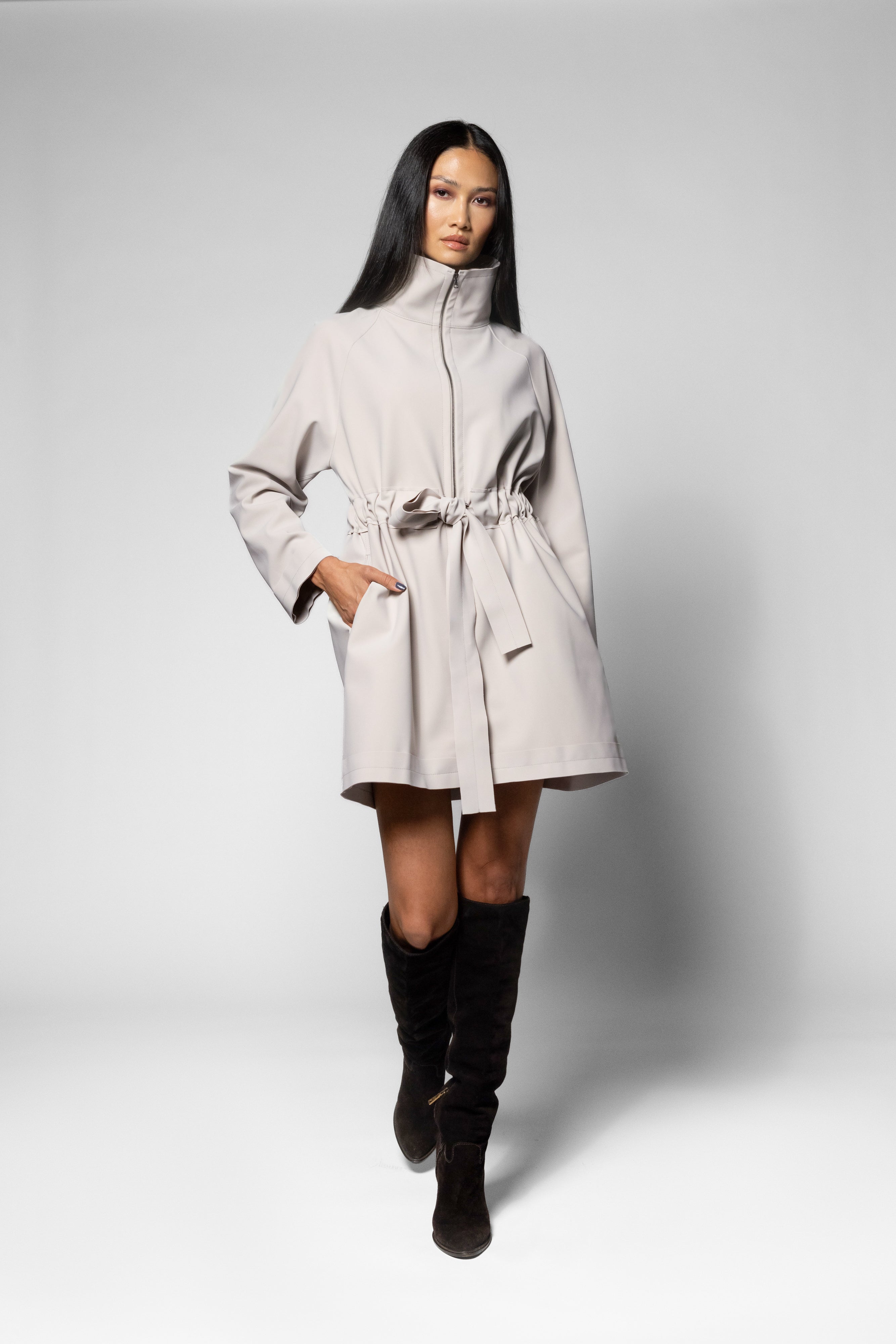 A woman modeling a light beige high-collar coat with a tie belt, paired with black knee-high boots, against a neutral gray studio background.