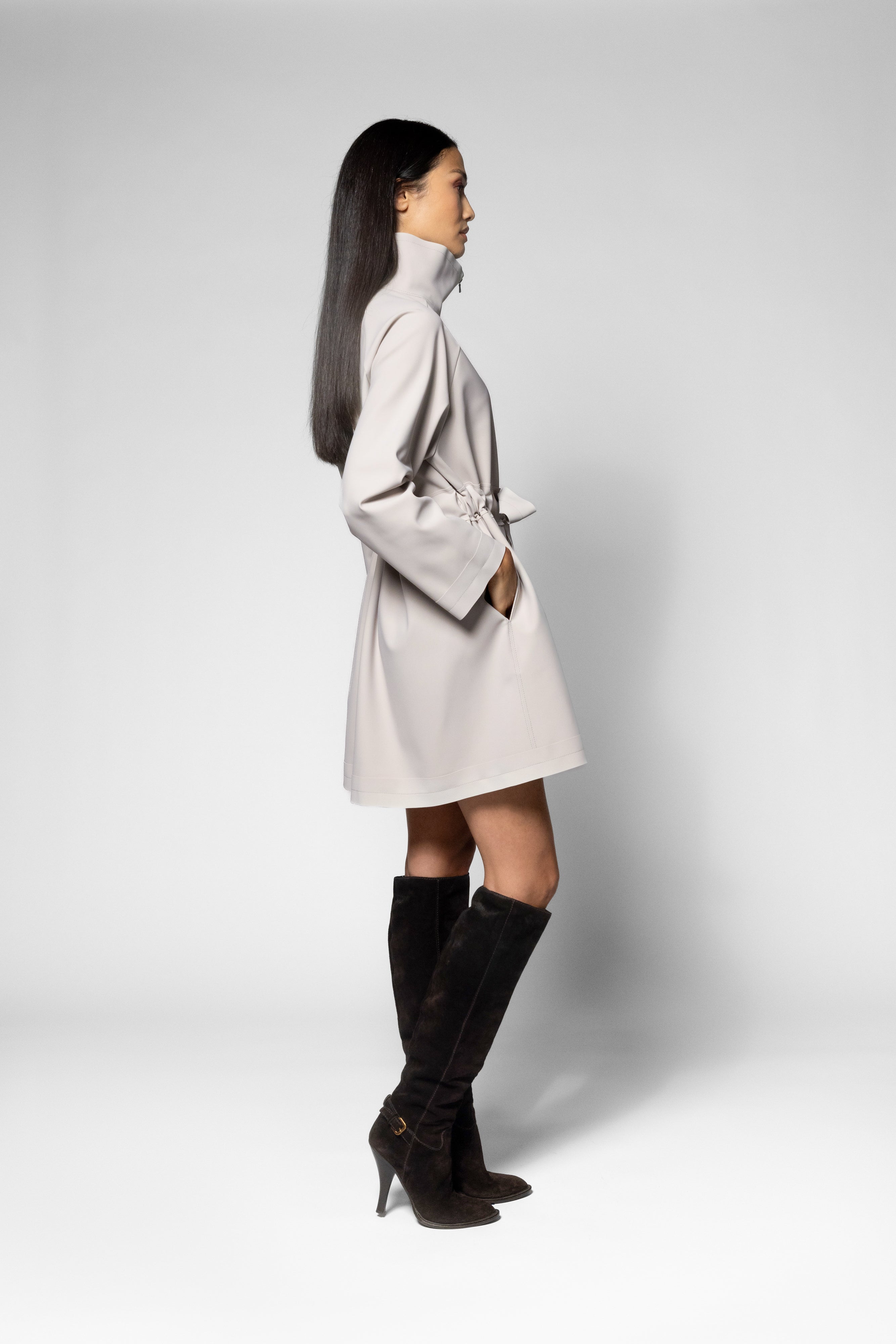 A woman in profile wearing a light beige wrap-style coat with a high collar and long sleeves, paired with black knee-high heeled boots, photographed against a light gray studio background.