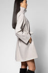 A woman shown in side profile wearing a light beige trench coat with a high collar and tie belt, paired with black boots, against a plain light gray studio background.