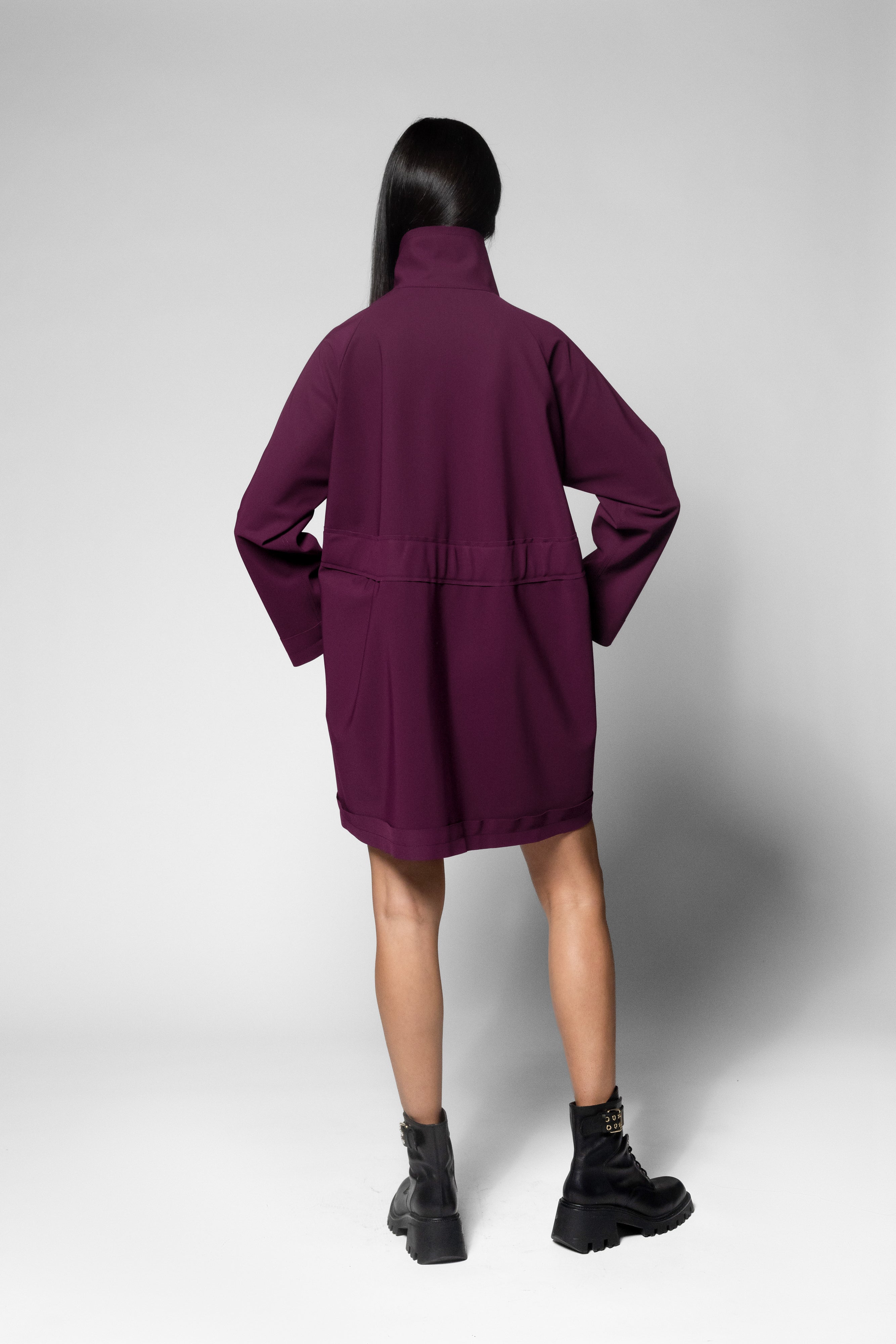 A model shown from the back wearing an oversized dark purple utility coat with a high collar and black combat boots, set against a light gray studio background.