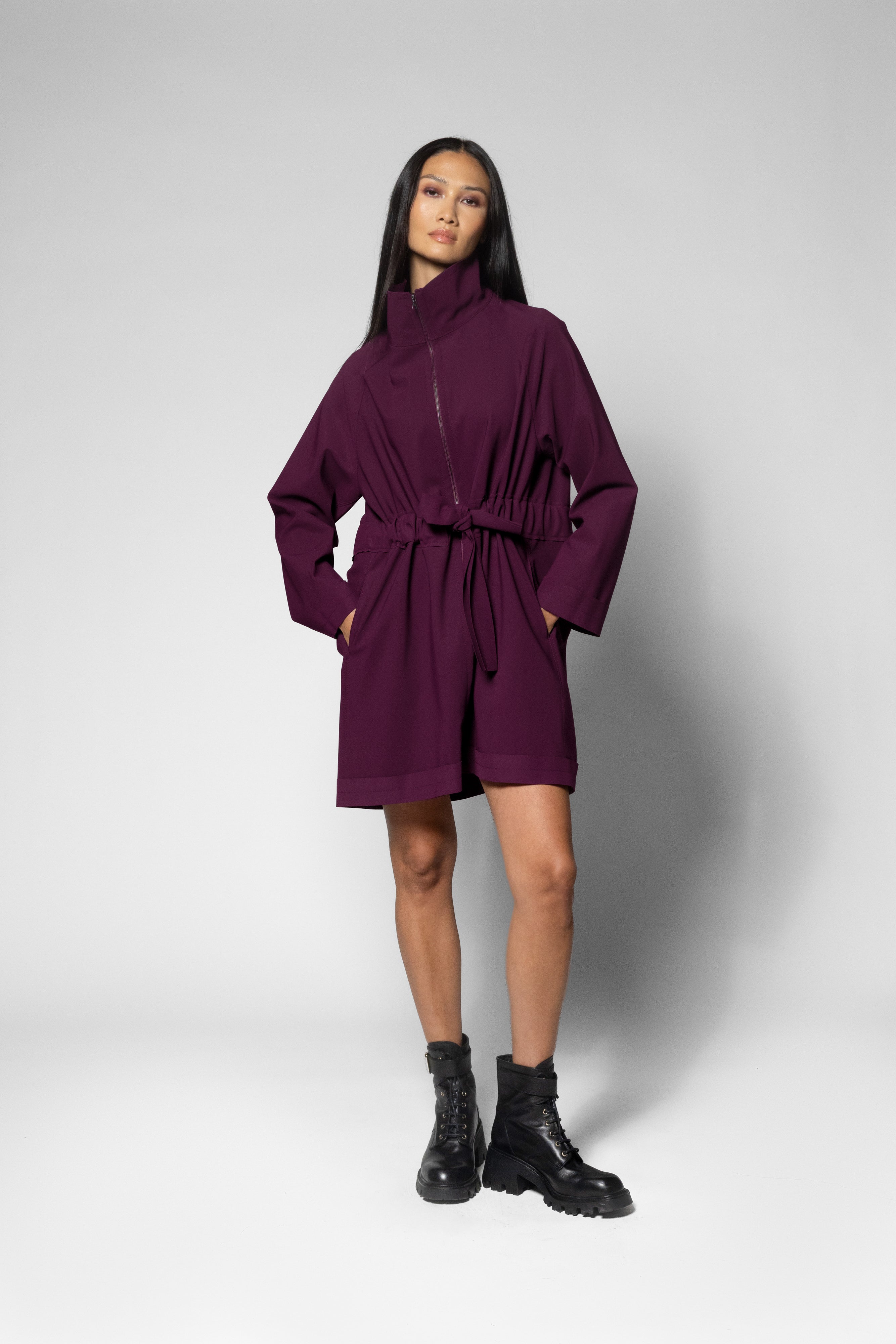 A model wearing a dark purple high-collar belted coat with long sleeves, paired with black combat boots, standing against a light gray studio background.
