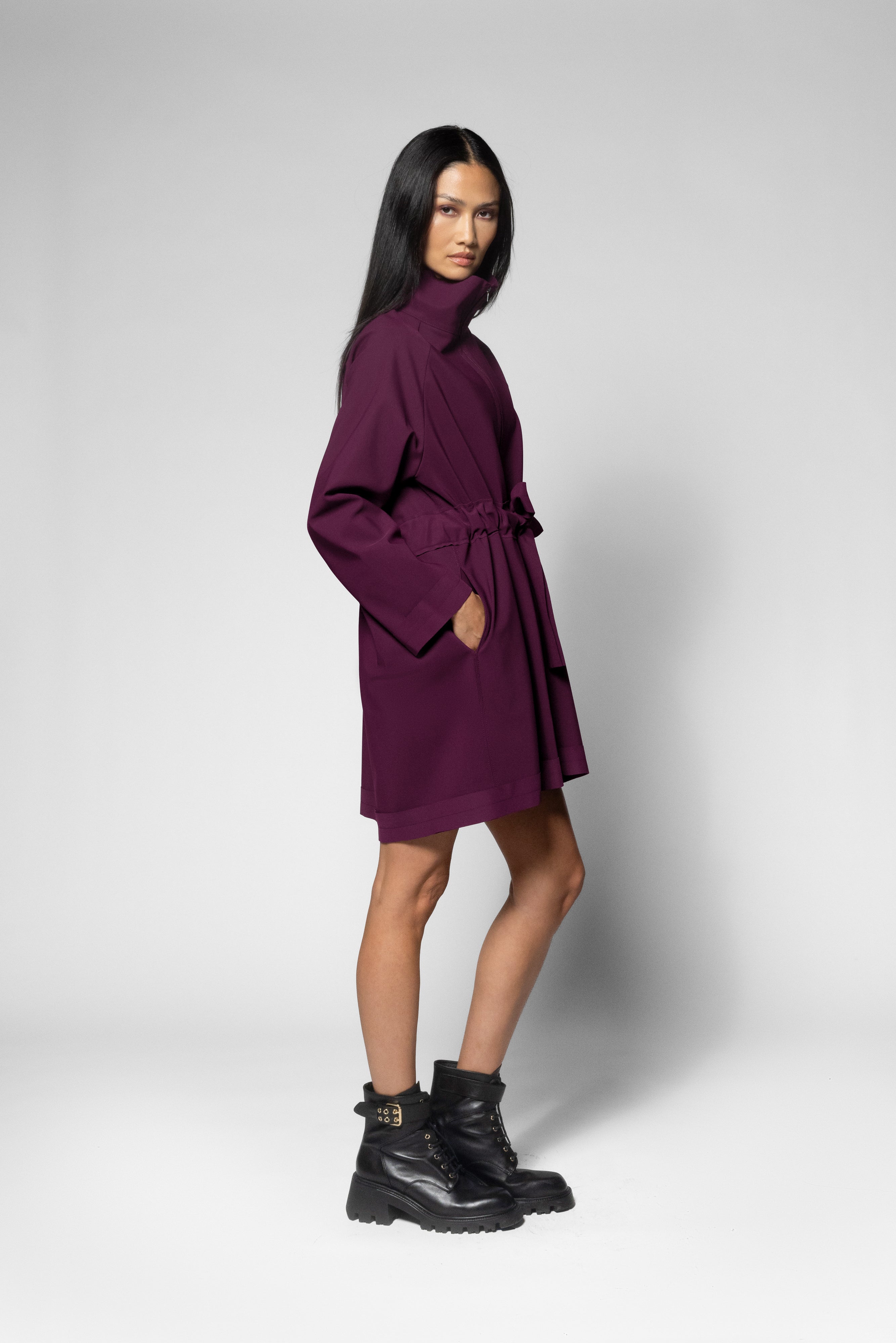 A model wearing a dark purple high-neck wrap coat with a tie belt, paired with black combat boots, standing against a light gray studio background.