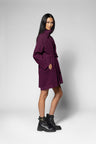 A model wearing a dark purple high-neck wrap coat with a tie belt, paired with black combat boots, standing against a light gray studio background.