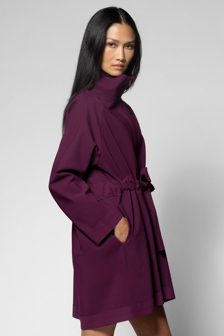 A woman modeling a long-sleeved, high-neck wrap coat in deep plum purple, featuring a self-tie belt at the waist and side pockets, set against a plain light gray studio background.