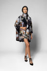 A model stands against a light gray studio background wearing a long-sleeved, high-neck wrap coat with a black base and a detailed multicolor floral and butterfly print. The coat features a large collar, wide sleeves, and a front flap.