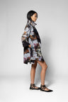 A model wearing a long-sleeved, knee-length trench coat with a dark base and a large, painterly floral pattern in pink, white, and brown, paired with black lace-up flat sandals against a light gray studio background.