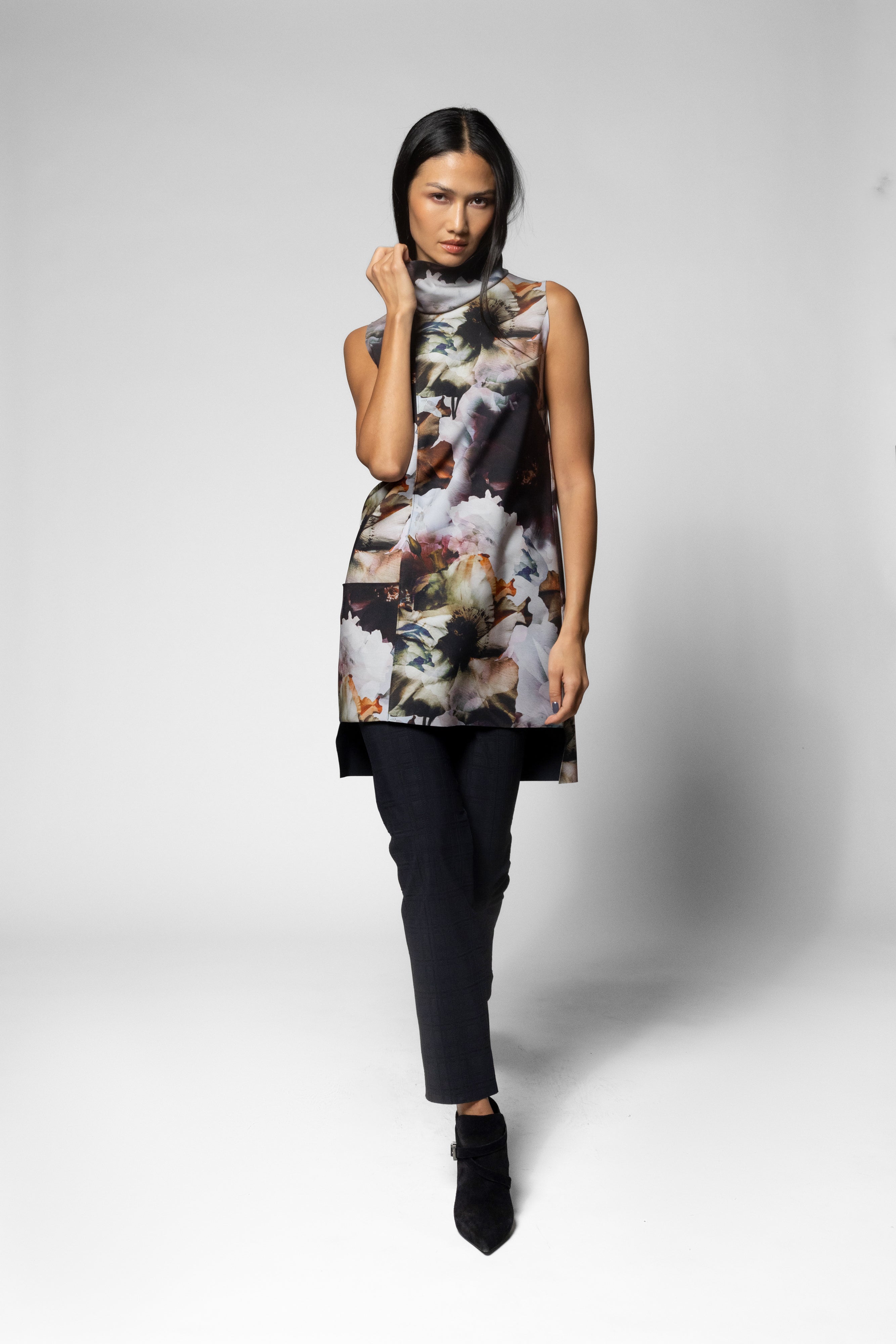 A model wearing a sleeveless mock-neck tunic top with a dark abstract floral print, paired with black pants and black pointed-toe ankle boots, against a light gray studio background.