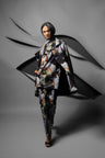 A model walks forward in a dramatic studio setting wearing a matching printed jacket and trousers set with black draped fabric elements behind them. The garment features an abstract floral or painterly pattern in black, gray, white, and muted orange-brown tones.