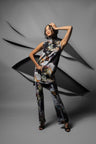 A model poses in a sleeveless high-neck tunic and matching wide-leg trousers set featuring a dark abstract floral print with orange, yellow, and white accents, set against a gray studio backdrop with a dynamic black graphic overlay.