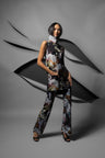 A model stands in a studio wearing a sleeveless high-neck tunic top and matching wide-leg pants in a dark floral and abstract print. The outfit features a mix of black, gray, white, and muted green tones.
