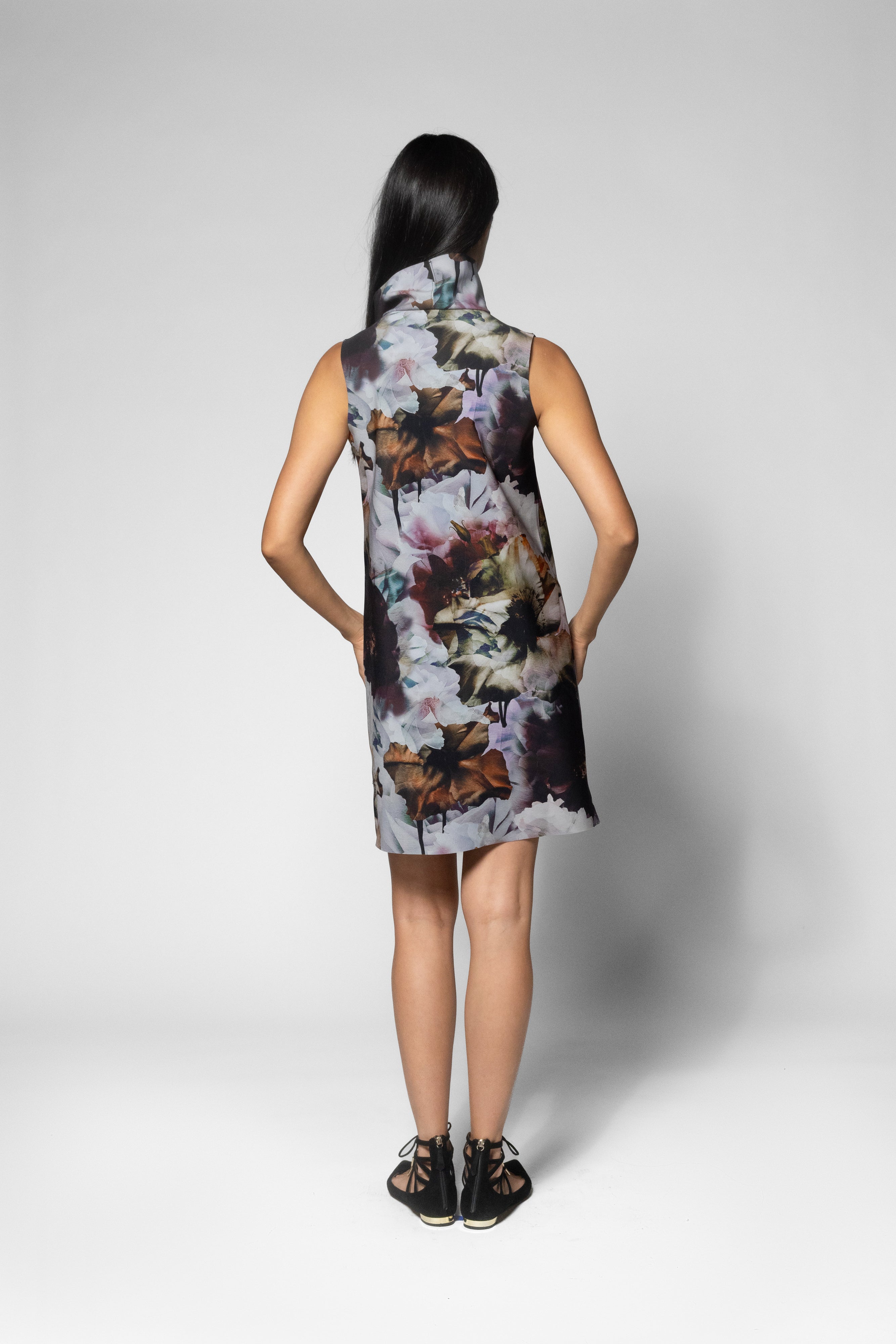 A woman viewed from the back wearing a sleeveless high-neck dress with a multicolor abstract floral print, standing against a light gray studio background.